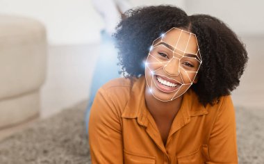 Yüz tanıma teknolojisi yüz hatlarını incelerken kıvırcık saçlı Afro-Amerikalı kadın gülümsüyor. Bu sahne modern tanımlama yöntemlerinin gelişigüzel bir ortamda entegrasyonunu yakalar..