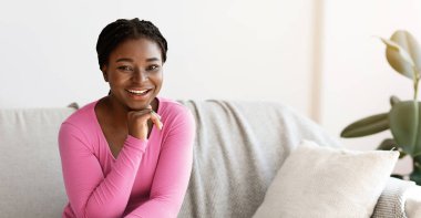 Pozitif genç Afro-Amerikalı kadın evdeki kanepede poz veriyor, rahat bir oturma odasında rahat bir koltukta dinleniyor, çenesini uzatıyor, kameraya gülümsüyor, hafta sonu eğlencesinin tadını çıkarıyor, boş alan arıyor.