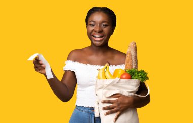 Neşeli genç Afro-Amerikalı kadın elinde süpermarket faturası ve market poşeti tutarken başarılı bir alışveriş, gıda bütçesi planlaması, sarı arka planda duran organik meyve ve sebzeler satın aldı.