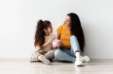 Family Savings. Happy Little Girl And Her Mom Holding Piggybank And Smiling While Relaxing On Floor Near White Wall At Home, Young Middle Eastern Mother Teaching Daughter Economy, Copy Space