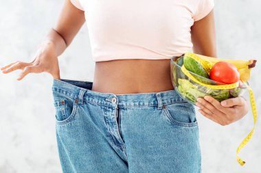 Zayıflatıcı diyet. Afro-Amerikalı bir kadın, elinde meyve, sebze ve açık arkaplanda bol bol kot pantolon giyer. Genç siyahi bayan kilo vermede başarılı oluyor.