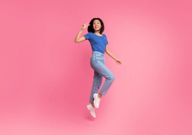 Mutluluk Konsepti. Genç Asyalı enerjik bir kadının tam boy portresi atlıyor ve pembe stüdyo arka planında iki parmak izole edilmiş zafer işareti işareti gösteriyor. Heyecanlı kadın zaferi kutluyor