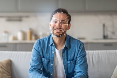 Bir adam, kimliğini doğrulamak için yüz tanıma teknolojisinin kullanıldığı çağdaş bir mutfakta koltukta otururken gülümsüyor. Modern tasarım teknolojide yeni bir normalliği yansıtıyor..