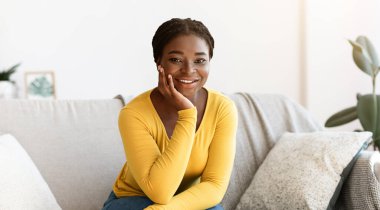 Huzur içinde yat. Oturma odasındaki koltukta dinlenen gülümseyen Afrikalı Amerikalı bir kadının portresi, kameraya bakan siyah bir kadının ev içi rahatlığının ve hafta sonu tatilinin keyfini çıkarması.