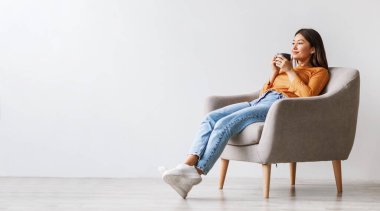 Milenyum Asyalı rahat bir kadın kahve içiyor, mola veriyor, beyaz stüdyo duvarına yaslanmış koltukta dinleniyor, boş alanı olan bir panorama. Hoş bir genç bayan serinliyor ve sıcak içkinin tadını çıkarıyor.