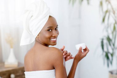 Yaşlanma önleyici vücut bakımı, nemlendirici ve tecrit sırasında evde kalmak. Genç, mutlu, Afro-Amerikan bayan blogcu beyaz havluyla yatak odasının içinde krem tutuyor ve sabahları kameraya bakıyor.
