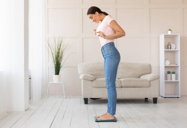 Kilo kaybı konsepti. Kilo tartısı üzerinde duran kız evde zayıf parmaklarını çapraz yaparak tartıyor. Boşluğu Kopyala