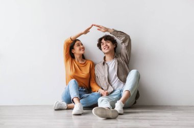 Milenyum Asyalı çiftin evlerinin çatısını elleriyle yapmaları, yeni boş bir dairede yerde oturmaları, beyaz stüdyo duvarına karşı poz vermeleri, tam boy portre. Taşınma günü, taşınma konsepti