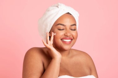 Mutlu siyah kadın yüzüne güzellik ürünü sürüyor ve gülümsüyor. Havluya sarılmış kadın duş üstüne yüz kremi kullanıyor. Pembe stüdyo arka planında izole edilmiş.