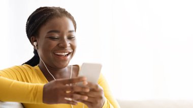 En sevdiği eğlence yeri. Neşeli siyah kadın evde akıllı telefon kullanıyor ve kulaklıkla müzik dinliyor, neşeli Afrikalı Amerikalı kadın oturma odasında koltukta dinlenirken internete bakıyor, boşluğu kopyalıyor.