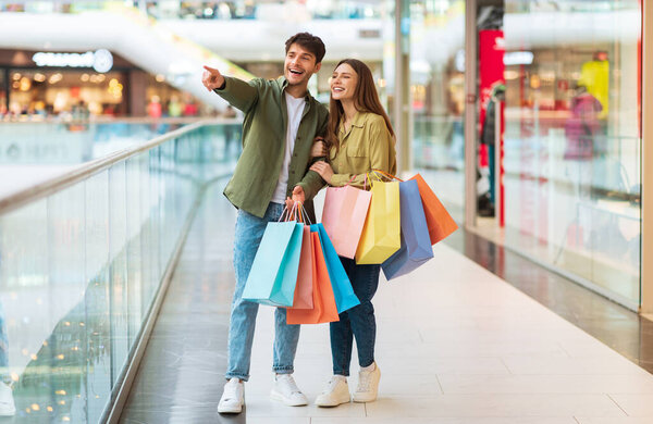 Веселые покупатели пара с красочные сумки покупки вместе указывая пальцем рядом стоять в современном торговом центре в помещении на выходные. Предложение сезона продаж и концепция шопаголизма. Полная длина