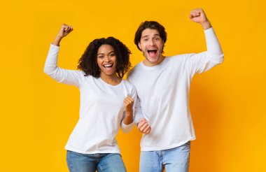 Yes. Overjoyed interracial couple celebrating success with raised fists, posing together over yellow background with empty space
