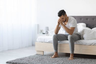 Despair Concept. Portrait Of Depressed Young Arab Man Sitting On Bed At Home, Upset Middle Eastern Man Leaning Head On Hands, Having Life Problems, Feeling Heartbroken And Lonely, Copy Space