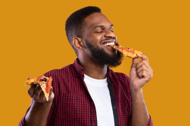 Siyah adam pizza yiyor. Sarı arka planda dikilen, gözleri kapalı ıvır zıvırın tadını çıkarıyor. Stüdyo çekimi. Abur cubur yeme sorunu ve sağlıksız beslenme kavramı