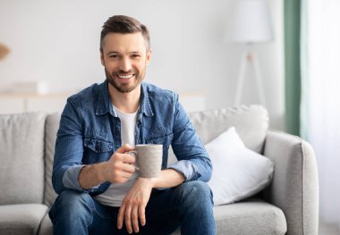 Bir fincan çayla kanepede oturan orta yaşlı, rahat oturma odasında kahve içen, kameraya gülümseyen, uzay fotokopisi çeken, beyaz sakallı bir adam.