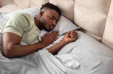 İyi uykular. Afrika kökenli Amerikalı bir adam yatak odasındaki rahat bir yatakta rahat bir şekilde uyuyor. İçeride gözleri kapalı dinlenen genç bir adam. Yurt içi rahatlama ve uyku zamanı kavramı