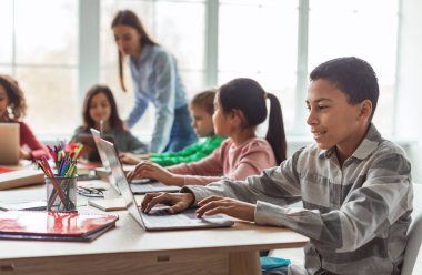 Cheerful Black Schoolboy Using Laptop Computer Learning Online With Diverse Classmates Sitting At Desk In Modern Classroom At School. E-Learning, Technology And Education Concept