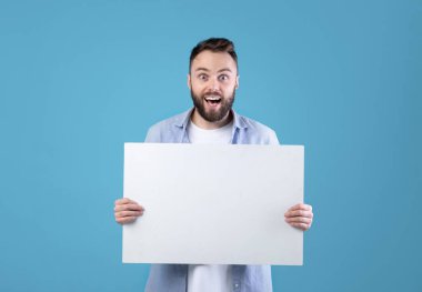 Heyecanlı genç adam elinde mavi stüdyo arka planında fotokopi alanı olan boş bir poster tutuyor. Mutlu milenyum adamı reklam tasarımınız için boş afiş gösteriyor.