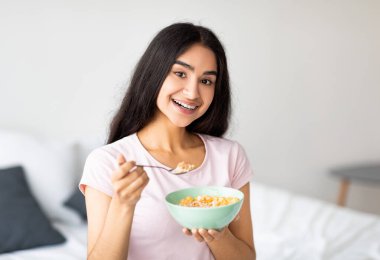 Lezzetli mısır gevreğini sütle yiyen, yatak odasında besleyici mısır gevreğinin tadını çıkaran hoş Hintli kadın. Genç Asyalı kadın dengeli kilo verme diyeti yapıyor, sağlıklı yemek yiyor.