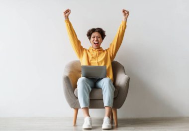 Duygusal genç Asyalı adam dizüstü bilgisayarla koltukta oturuyor, heyecan içinde ellerini kaldırıyor, beyaz stüdyo duvarına karşı başarıyı kutluyor, tam boy. Millennial Man online satışın tadını çıkarıyor ya da büyük bir zafer