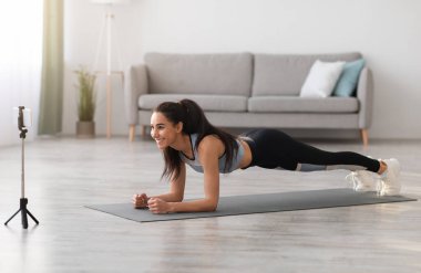 Spor giyinen neşeli genç bayan oturma odasında planking yapıyor, fitness videosu izliyor ya da spor eğitmeniyle video konferansı yapıyor spor yapan kadın spor yapıyor ve akıllı telefon ekranına bakıyor, boş alan var.
