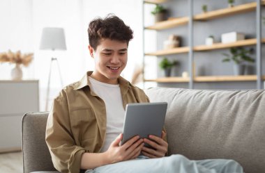 People And Technology. Portrait Of Smiling Asian Man Holding And Using Digital Tablet Sitting On Couch Indoors In Living Room. Happy Male Surfing Internet, Watching Movie Online