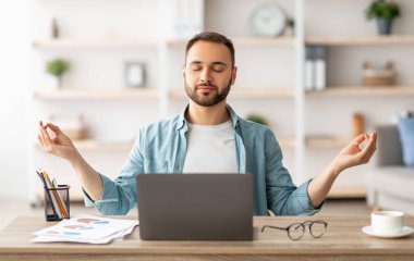 İş yeri stres yönetimi. Sakin Kafkasyalı adam ofisteki bilgisayarın önünde gözleri kapalı meditasyon yapıyor. Genç erkek serbest çalışan, kendini huzurlu ve dengeli hissediyor, masasında yoga yapıyor.