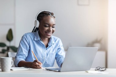 Siyah kadın ofis çalışanı iş yerinde dizüstü bilgisayarda giyinen ve not alan, kulaklık takan, boş alan arayan.