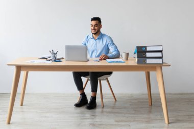 Modern ofiste dizüstü bilgisayarda çalışan gülümseyen Orta Doğulu erkek menajerin portresi, masada oturan ve bilgisayarını kullanan neşeli adam, ekranda Webinar 'ı izleyen, bedava fotokopi çeken,