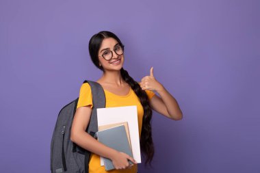 Eğitim Konsepti. Gözlüklü gülümseyen Hintli kız öğrencinin portresi işaret işareti gösteren, elinde kameraya bakan defterler tutan. Sırt çantalı bir kadın, mor stüdyo geçmişi var.