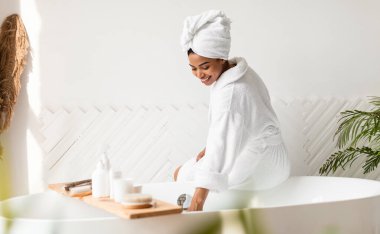 Sıhhat ve rahatlama. Mutlu Afrikalı Amerikalı Kadın Suya Dokunma Banyo Hazırlıyor Evdeki banyoda, üzerinde bornozuyla. Güzellik Bakımı Rutin Konsepti