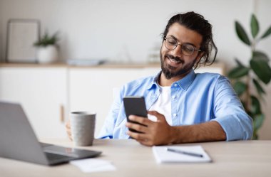 Sırıtkan Hintli çalışanlar akıllı telefon kullanıyor, ofiste kahve içiyor, işte tatilin tadını çıkarıyor, arkadaşlarla mesajlaşıyor ya da sosyal ağlara takılıyor, dizüstü bilgisayarla masada oturuyor, seçici odak noktasıyla boş alan arıyor.