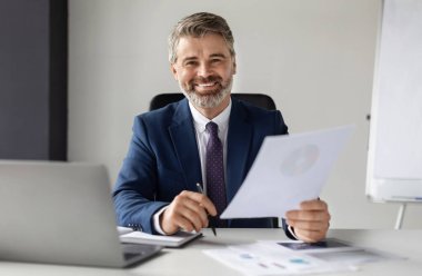 Ofisteki belgelerle çalışan ve kameraya gülümseyen olgun bir iş adamı, orta yaşlı şirketin CEO 'su, mali raporları tutuyor, dizüstü bilgisayarla masada oturuyor, boşluğu kopyalıyor,