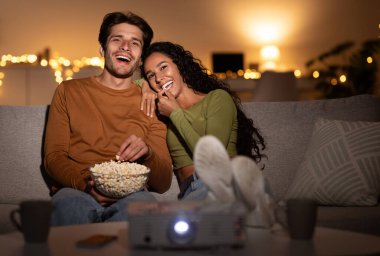 Ev sineması. Eşler komedi filmi izlerken güler, patlamış mısır yer ve geceleri koltukta oturur. Hafta sonu akşamı birlikte eğlenceli bir film izleyen genç çift. Ev eğlencesi ve eğlence.