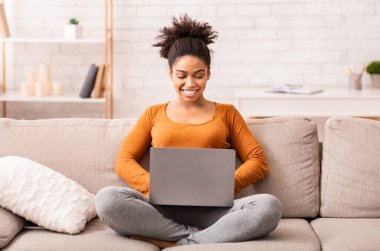 Serbest Yaşam Tarzı. Neşeli Afro Kadın dizüstü bilgisayarda çalışıyor ve internette evdeki koltukta oturuyor. Seçici Odaklanma