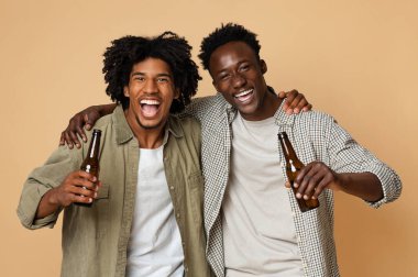 İki mutlu siyahi arkadaşın portresi, bira şişelerini kucaklayıp tutarken, neşeli Afrikalı Amerikalı erkeklerin bej arka planda birlikte poz verirken eğlenirken, Stüdyoda eğlenirken,