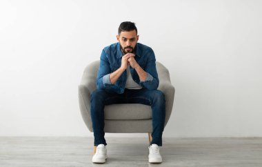 Olumsuz duygular. Genç bir Arap, kasvetli veya düşünceli bir yüz ifadesiyle koltukta oturuyor, mutsuz ya da yorgun hissediyor, problem yaşıyor, beyaz stüdyo duvarına karşı depresyon yaşıyor.