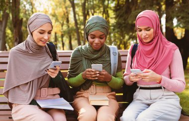 Dışarıdaki bankta oturan sosyal medyada cep telefonu mesajlaşması ve ağ kuran üç Müslüman kadın. İslami Öğrenci Kadınlar Akıllı Telefon ve Cep Telefonu Kullanıyor, Gadget Yaşam Tarzı Konsepti