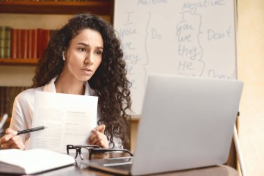 Uzaktan kumandalı çalışma. Kulaklık takmış ciddi bir kadının bilgisayarda video konferansı yapması, web kamerasına kalemle kağıt belge göstermesi, sunum yapması veya öğrencilere çevrimiçi sınıf vermesi.
