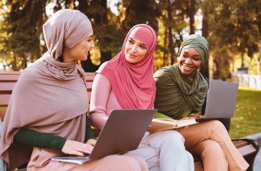 Laptopları ve kitaplarıyla birlikte parkta bankta oturup sohbet eden üç modern Isamic hanımefendisi. E-Öğrenim, Üniversite Öğrencileri Yaşam Tarzı, Müslüman Kadınlar İçin Eğitim
