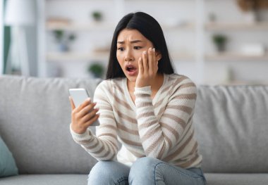 Oh hayır. Evde koltukta otururken akıllı telefondan kötü mesaj okuyan Asyalı kadın, ellerinde cep telefonuyla Koreli bir kadının rahatsız edici haberlere duygusal tepki göstermesi,