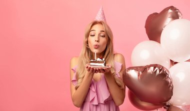 Doğum günü şapkalı ve balonlu hoş bir bayan pembe stüdyo arka planında mum üflüyor, fotokopi odası olan bir panorama. Milenyum kadını bayram ya da kutlama yapıyor.