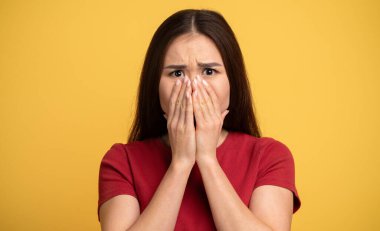 Asyalı genç kadın ağzını ve yüzünü elleriyle kapatarak kaygılarını dile getiriyor. Canlı sarı bir arkaplanın önünde duruyor, duygularını ve ifadesini vurguluyor..