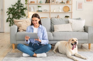 Evde vakit geçirmek. Elinde dijital tabletle halıda Golden Retriever 'ıyla oturan mutlu kadın portresi. Oturma odasında dinlenen bir bayan. Cihaz sörfü ağı ya da internetten alışveriş yapıyor.