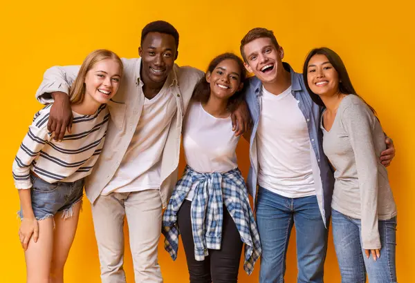 Sarı arka planda kucaklaşan ve gülümseyen çok ırklı öğrencilerden oluşan arkadaş canlısı bir grup.