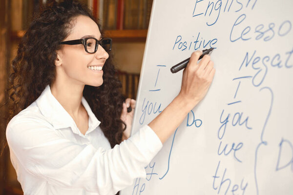 Education Cocnept. Smiling professional English teacher standing at whiteboard, writing with marker. Young female lecturer wearing glasses, looking at board, explaining grammar rules to the students