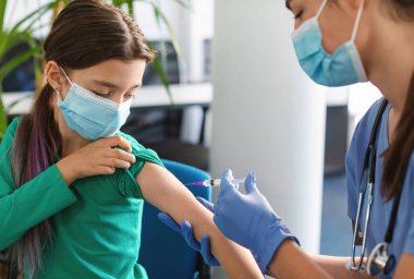Küresel salgın sırasında yeni Covid aşısı için klinik deney. Hastanede yüz maskesi takan küçük bir kız iğne oluyor. Doktor ya da hemşire şırıngayla kadın hastaya iğne yapıyor.