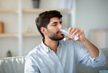 Genç Arap adam camdan tatlı su içiyor, oturma odasındaki kanepede oturuyor. Sağlık, diyet, evde düzgün beslenme, sabah ve ev yaşamında susuzluk, hidrasyon konsepti