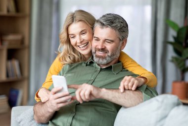 Mutlu orta yaşlı çift evde cep telefonunu birlikte kullanıyor internete ya da alışverişe bakıyor, kocası akıllı telefon ekranını işaret ediyor, gülümseyen karısına bir şey gösteriyor, yakın plan çekimleri yapıyor.