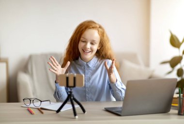 Blog Kavramı. Gülümseyen kızıl saçlı genç kız yaşam tarzı blogu kaydediyor tripodda cep telefonuyla konuşuyor, evdeki masada oturuyor. Kanalı için video bloğu çeken mutlu genç oyuncu.